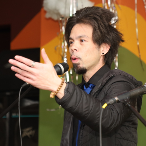 A man reads poetry at a microphone. Photo credit goes to ASTP Photography.