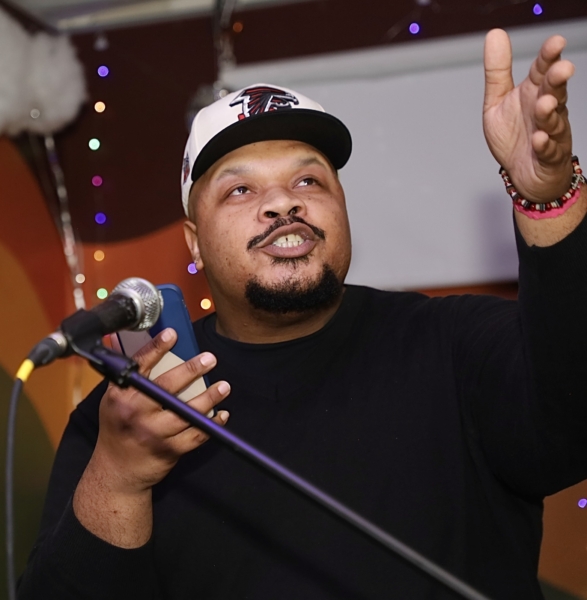 A man reads poetry at a microphone. There are cloud-like balloons in the background. Photo credit: ASTP Photography.